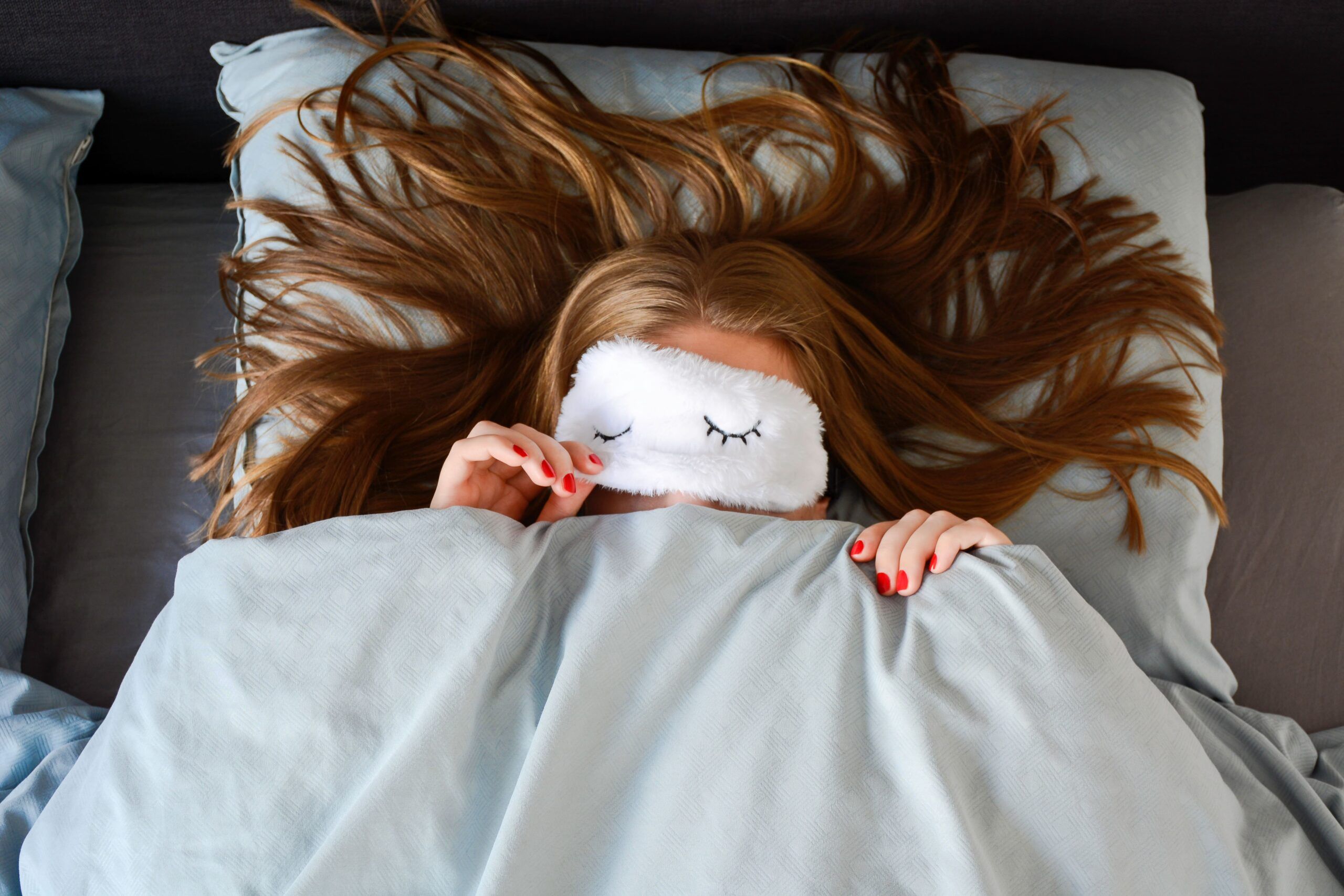 Junge-Frau-mit-langen-Haaren-schlaeft-im-Bett-mit-Schlafmaske-und-guckt-unter-der-Schlafmaske