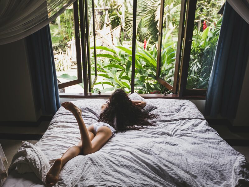 Frau in weißen Dessous faulenzt morgens im Bett, Blick aus dem Fenster auf tropischen Garten