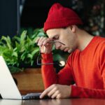 Müder Mann, der während der Online-Arbeit am Laptop eine Pause einlegt, Kopf in der Hand hält und Energiemangel verspürt.